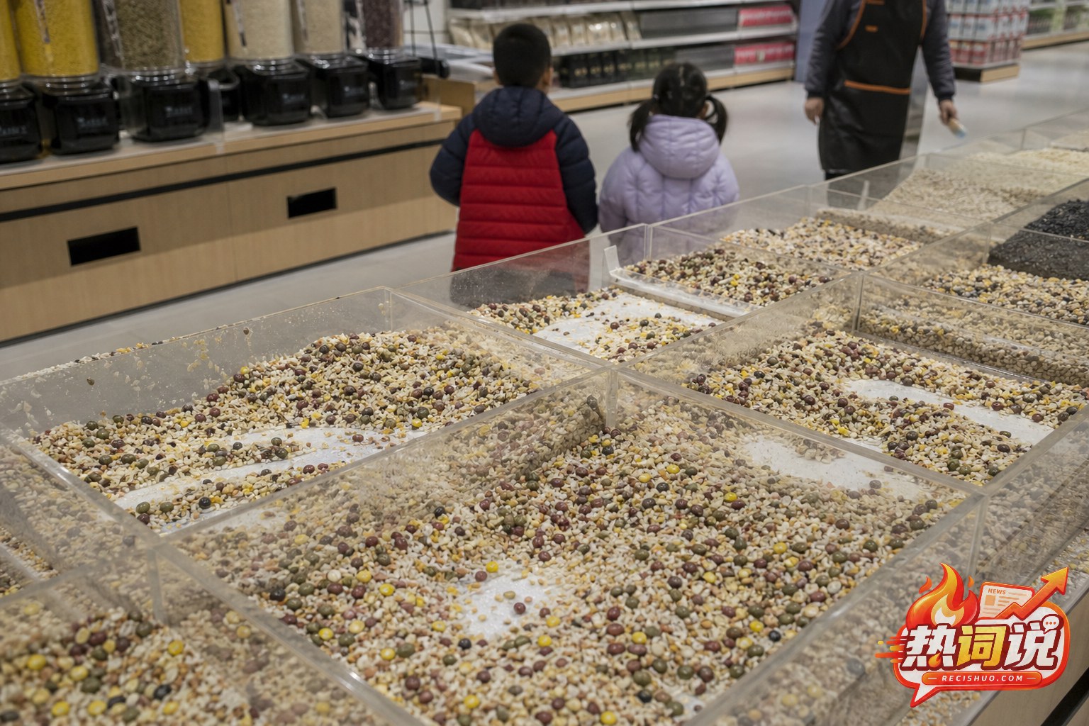超市杂粮区多个货柜内不同豆类和谷物混在一起,两名儿童背对镜头站在通道内