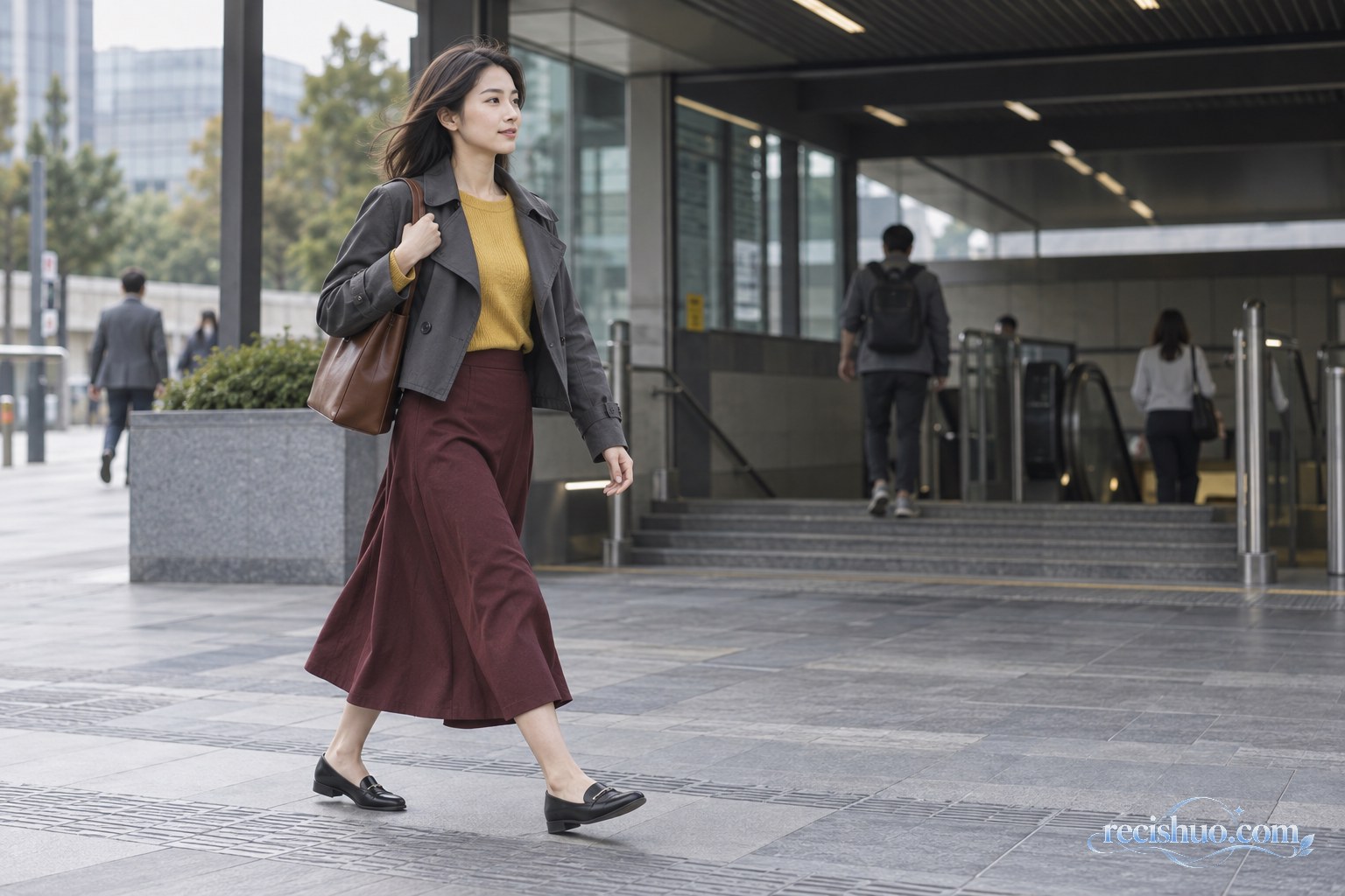 女性身穿芥末黄针织衫、深灰短风衣和酒红色半裙走过地铁站外