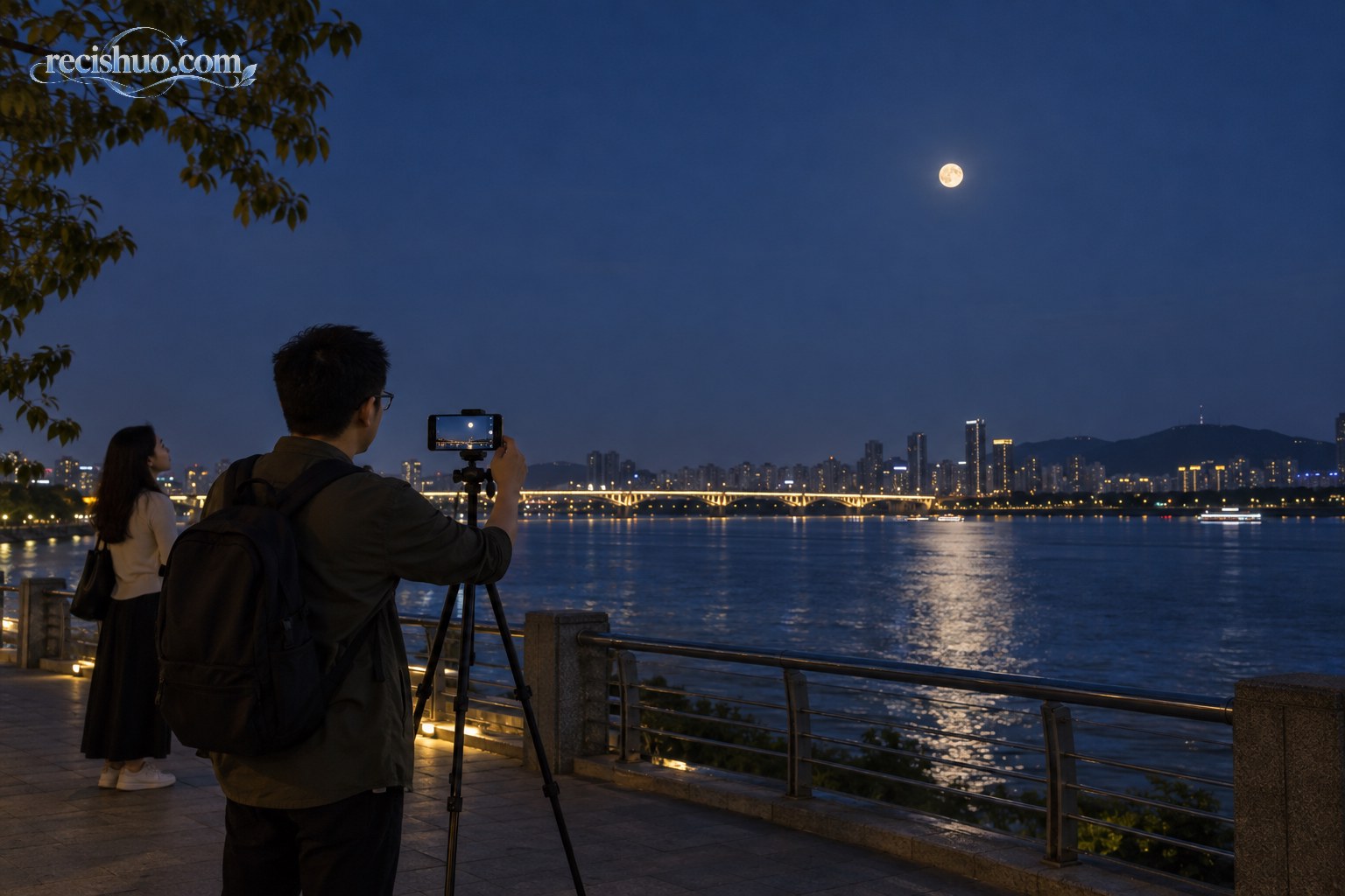 秋夜河畔,一名游客用三脚架固定手机拍摄远处的月亮