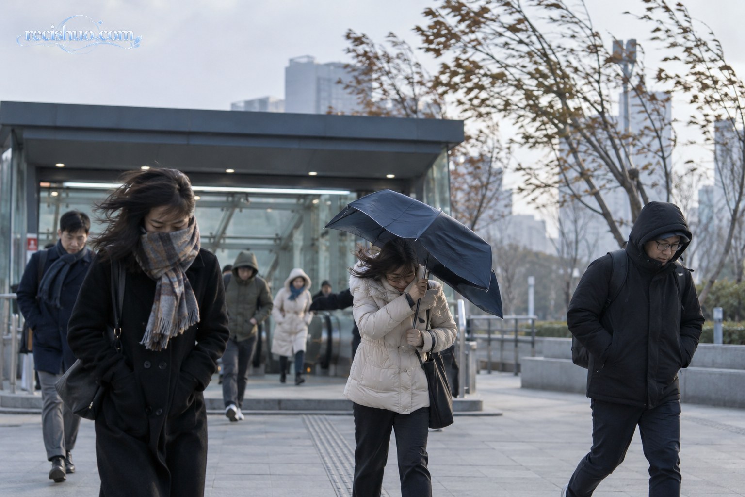 大风降温天气中,穿着保暖外套的通勤者在城市地铁入口附近迎风行走