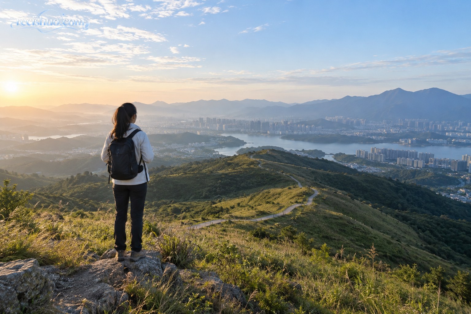 清晨,一名旅行者站在山坡步道上眺望远处的城市、湖泊与群山