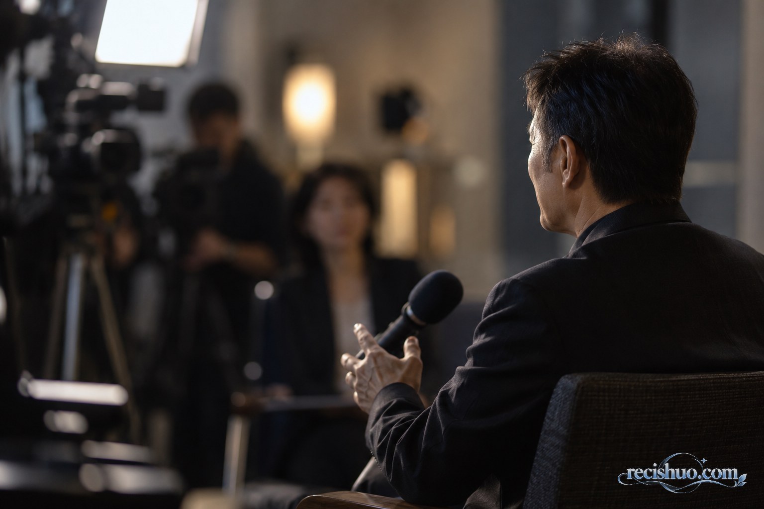 成熟华人男性公众人物在媒体采访空间接受访问的侧后方画面